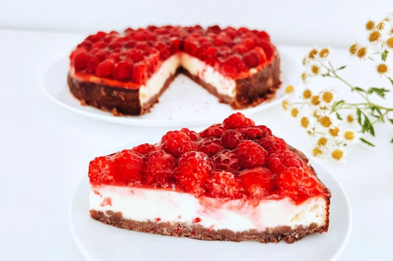 Kousek malinového cheesecaku s celým dortem na bílém podkladu.