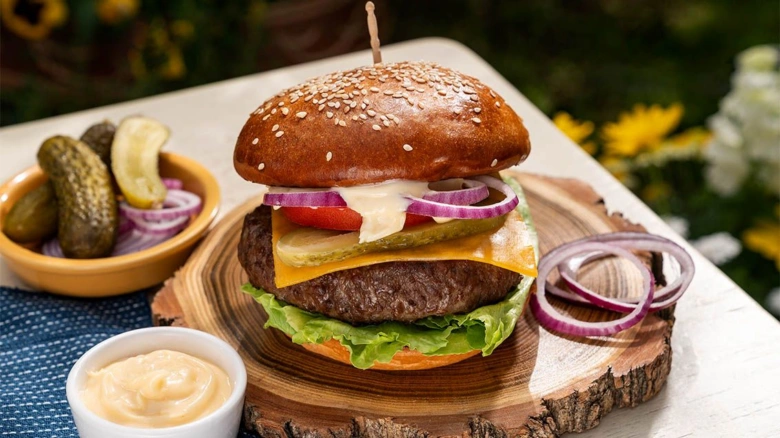 Domácí hovězí hamburger s sýrem, okurkou a cibulí na dřevěném talíři.