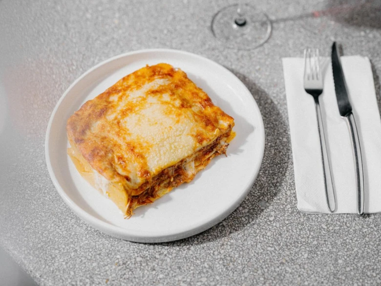 Kousek pečené lasagne na bílé talíři s vidličkou.