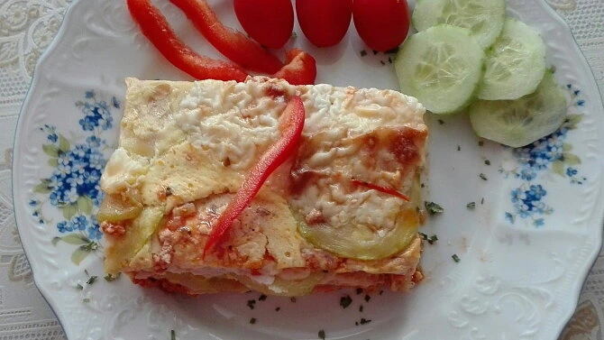 Kousek pečené cuketové lasagne s rajčaty a okurkou na talíři.