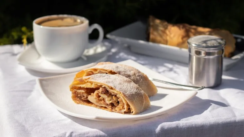 Kousek jablkového štrúdlu s kávou na bílém talíři.