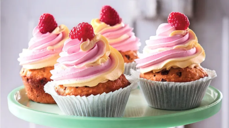 Čtyři muffiny s růžovou šlehanou smetanou a malinami na zeleném podstavci.