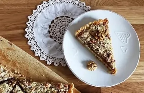 Kousek koláče s ořechy na bílém talíři vedle dřevěného prknka.