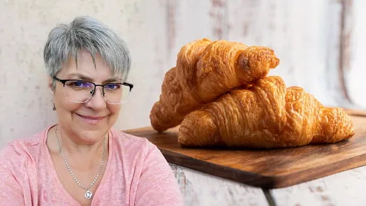 Žena s brýlemi stojí vedle dvou zlatých rohlíků na dřevěném prknu.