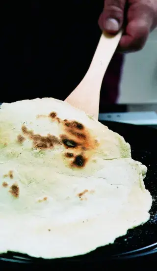 Ruce s dřevěnou lžící zvedají pečenou placku na pánvi.