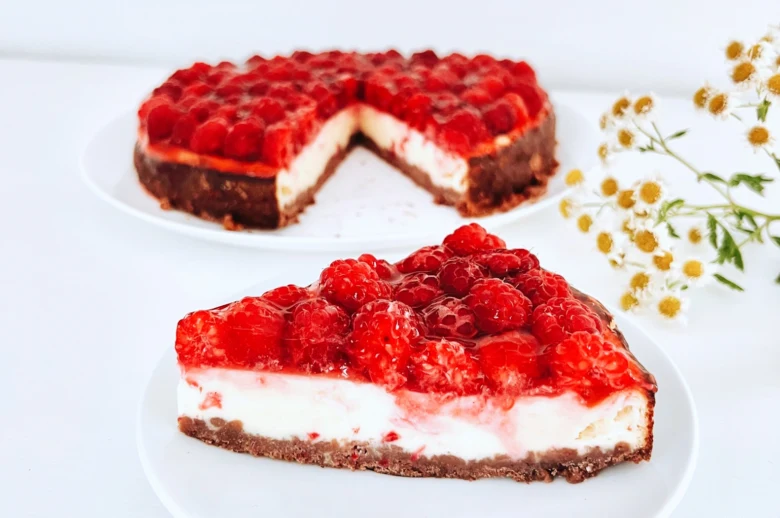Kousek malinového cheesecaku s celým dortem na bílém talíři.
