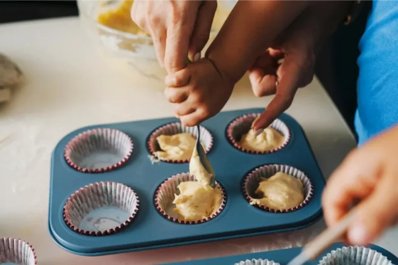 Dospělá ruka s dítětem nalévají těsto do formiček na muffiny.