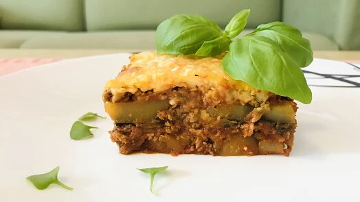Kousek zapečeného lilkového lasagne s bramborami na bílém talíři.