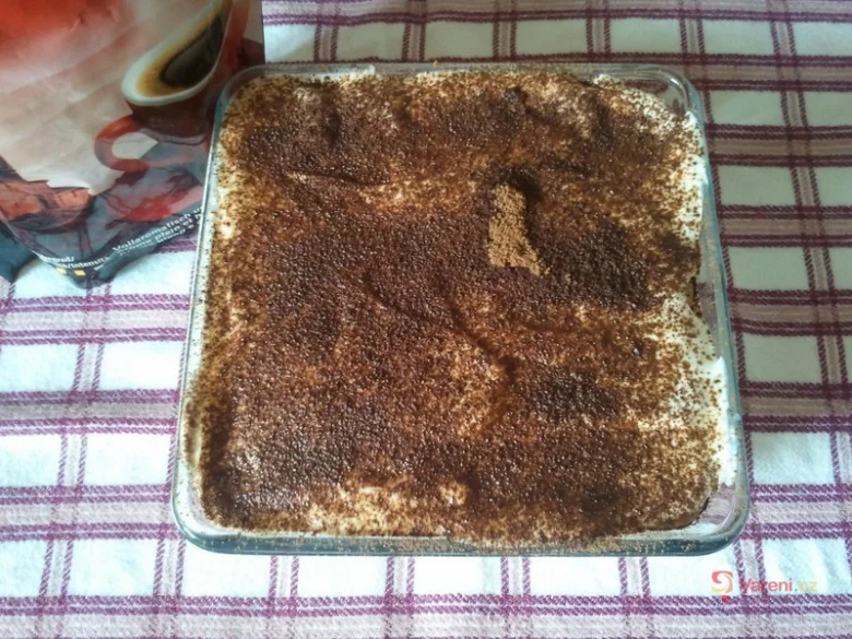 Tiramisu dezert v skleněné míse posypaný kakaem na pruhovaném ubrusu.