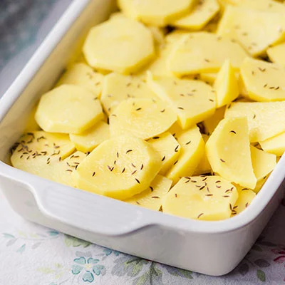 Nakrájené brambory s kořením v bílé pekáči připravené k zapékání.