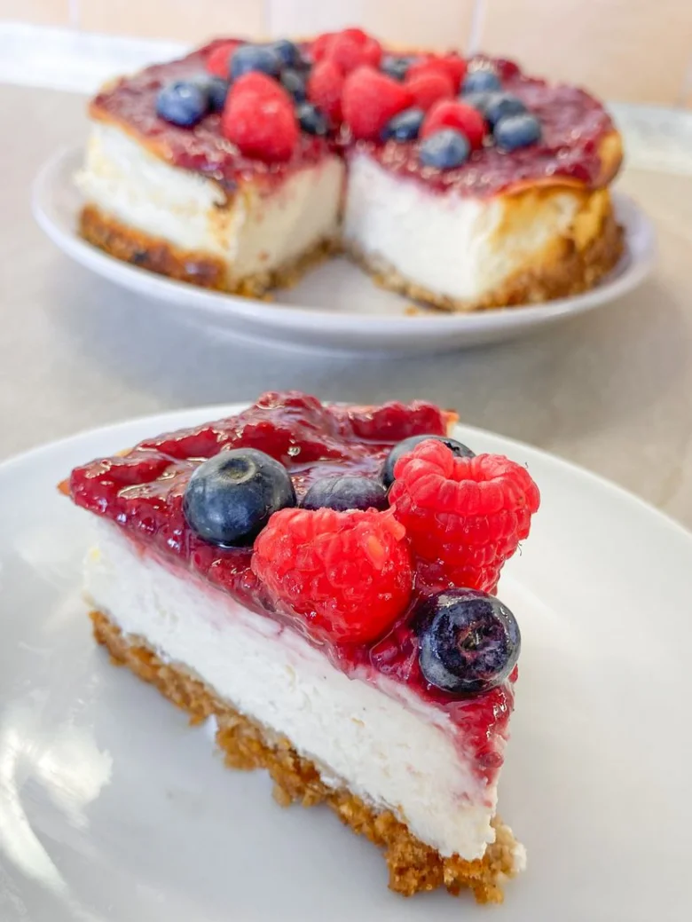 Kousek ovocného cheesecaku s malinami a borůvkami na bílém talíři.