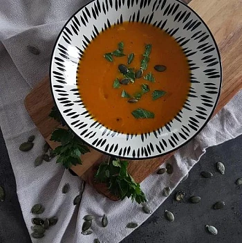 Mísa s oranžovou polévkou posypaná bylinkami na dřevěném podložce.